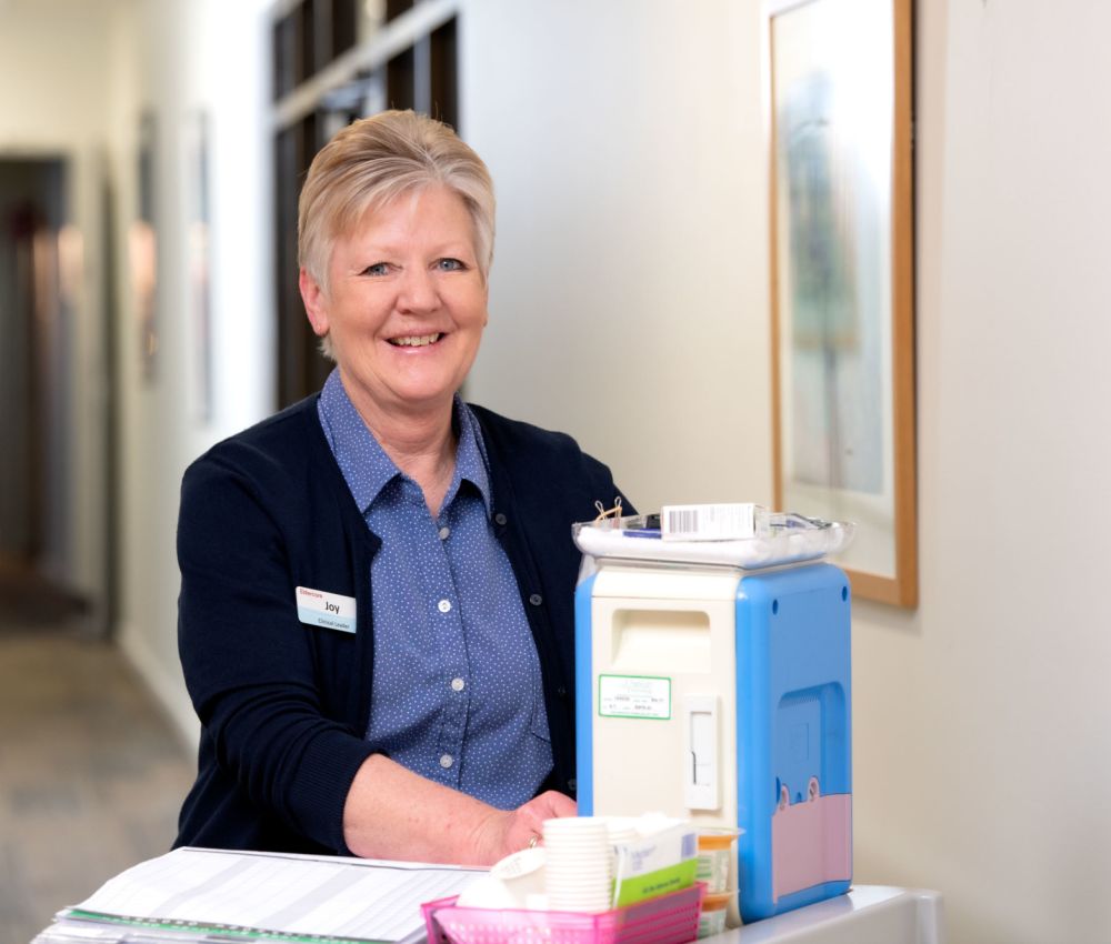Eldercare Clinical Leader Joy smiling at the camera from behind a medication trolley.