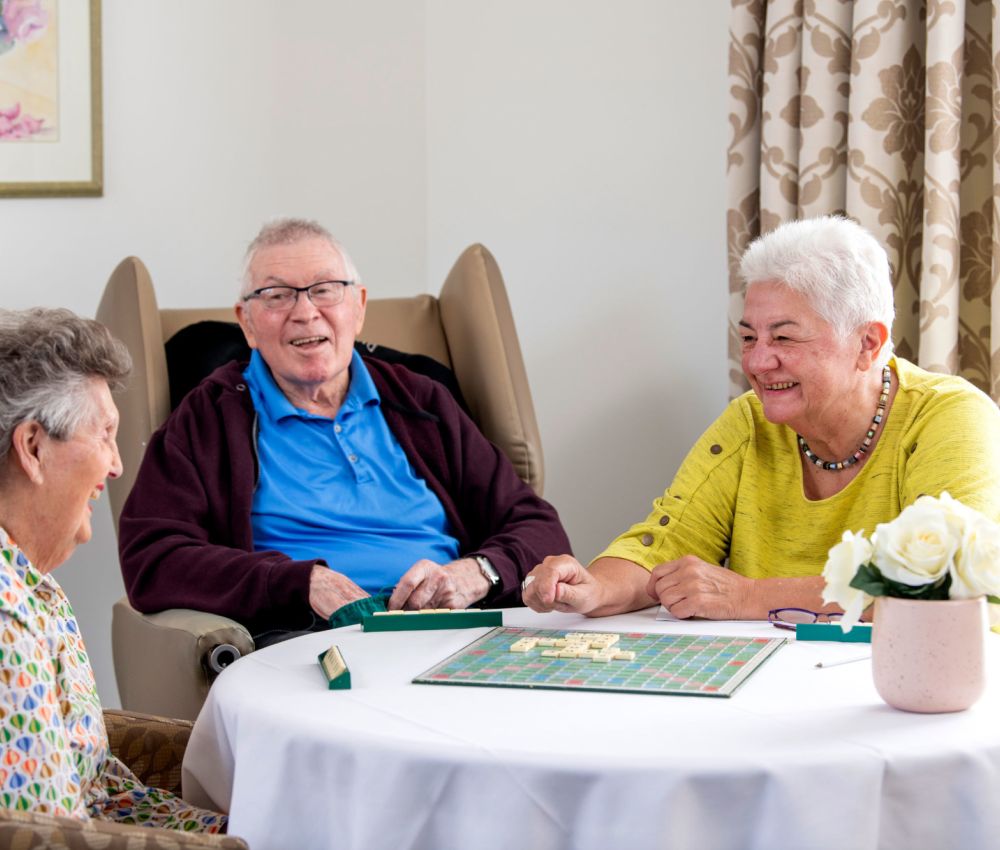 Eldercare The Lodge residents and volunteer playing Scrabble around a table and smiling at each other.