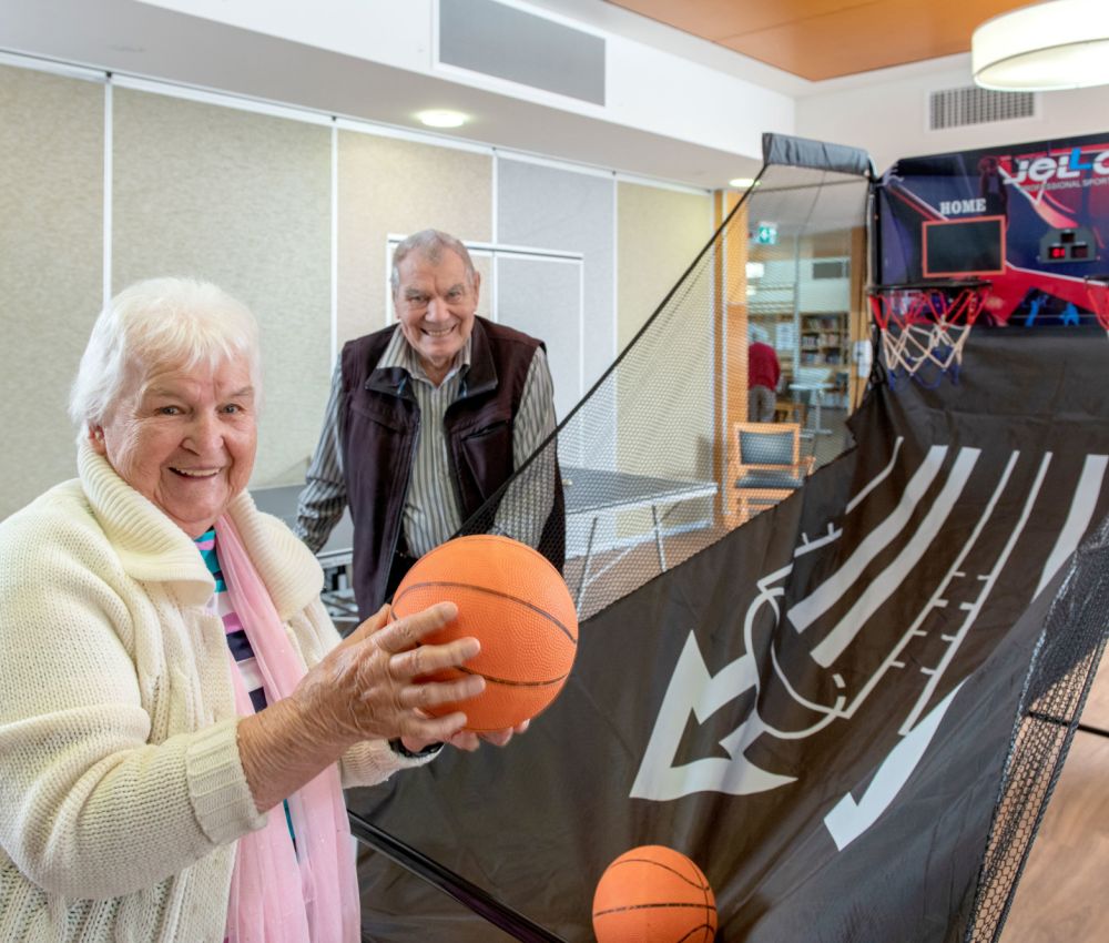 Eldercare Seaford residents Ruth and Otto smiling at the camera while Ruth plays with an arcade-style basketball game.