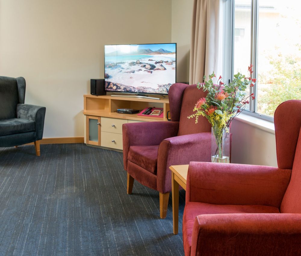 Lounge area at Eldercare Seaford complete with armchairs, television and a bunch of flowers in a vase.