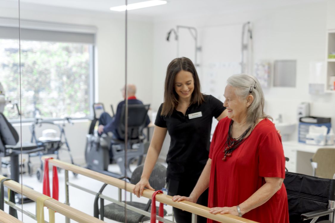 Eldercare physiotherapist Radhika Dick with Eldercare Allambi resident Bev Young at a set of parallel bars and smiling.