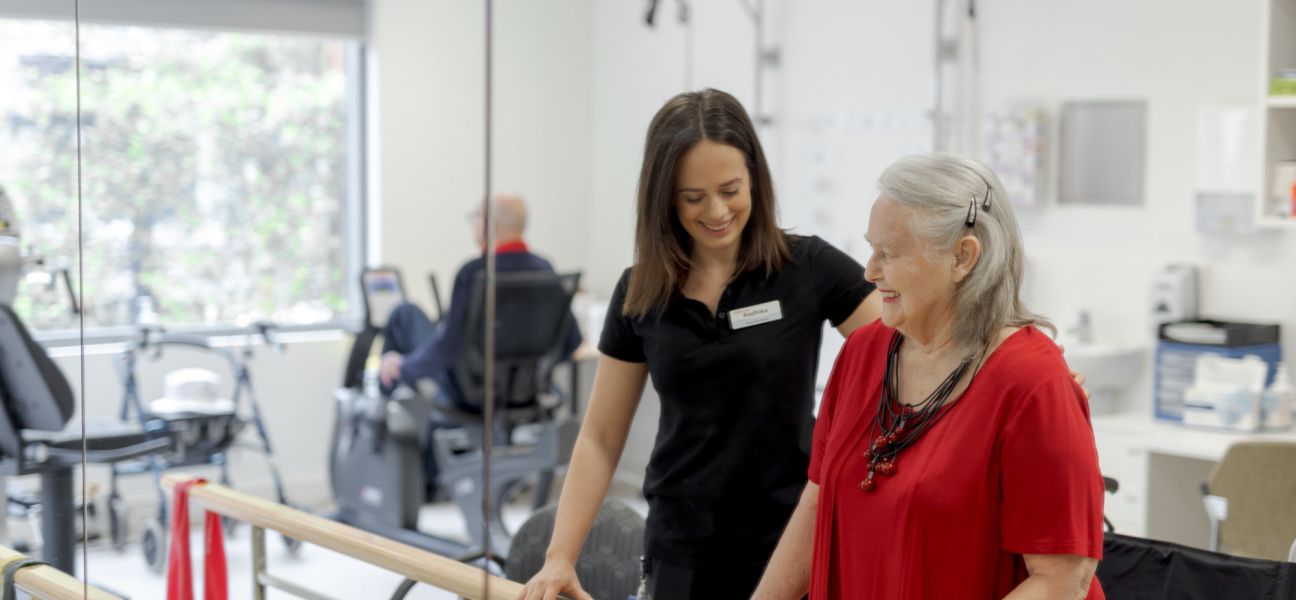 Eldercare physiotherapist Radhika Dick with Eldercare Allambi resident Bev Young at a set of parallel bars and smiling.