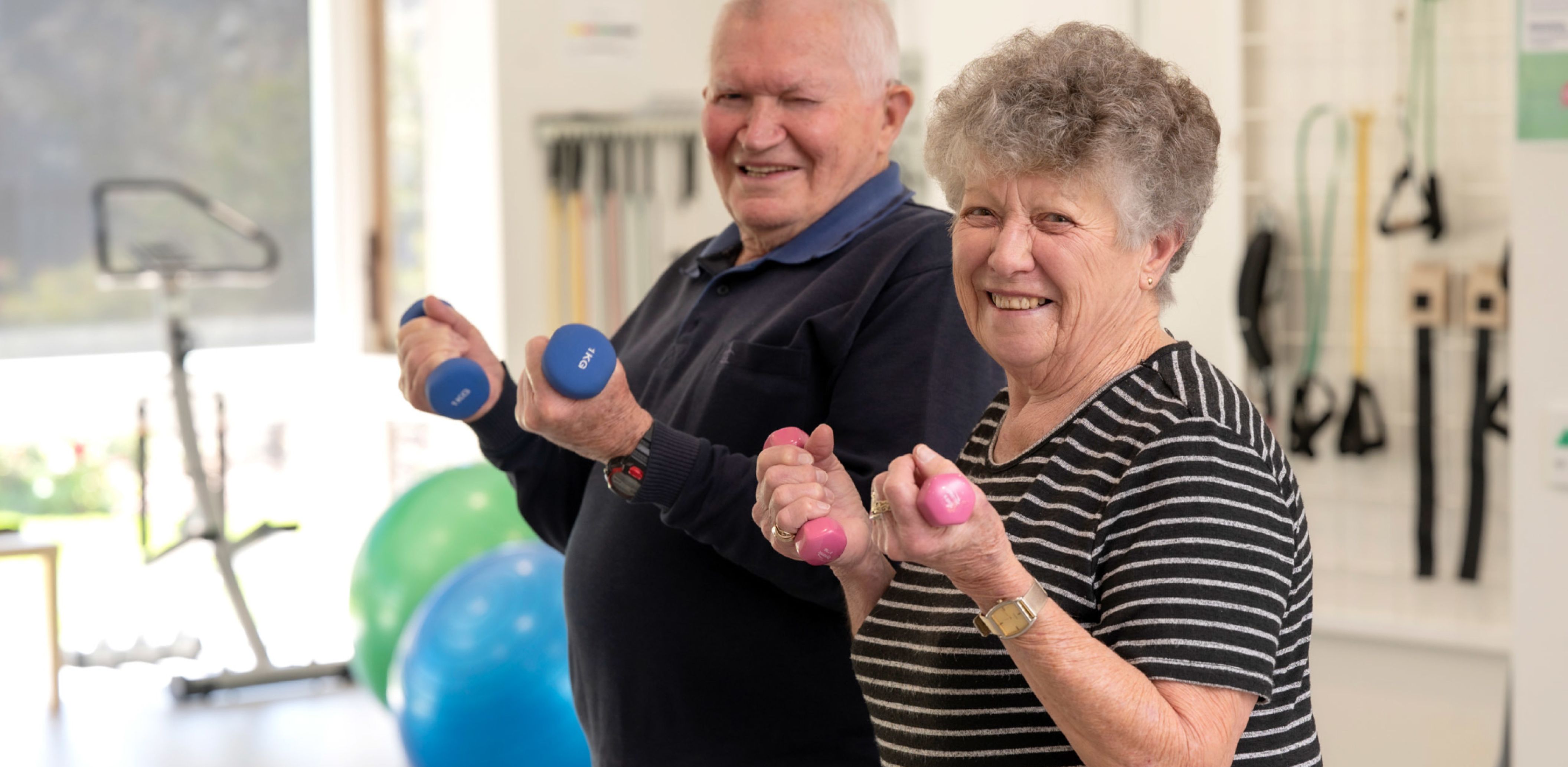 Two day therapy clients, a married couple, hold two dumbbells each and smile into the camera.