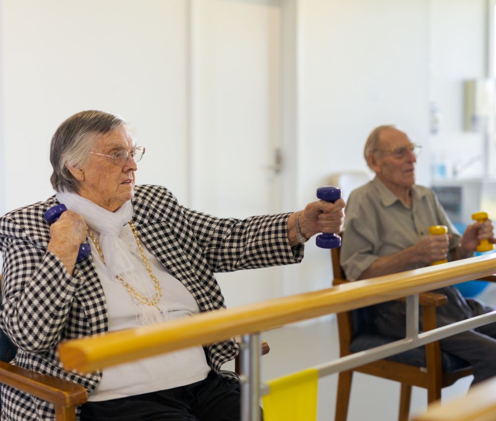 Two Eldercare residents sit at a set of parallel bars and use dumbbells to complete arm exercises.