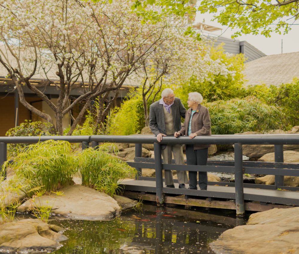 Two Eldercare residents, a couple, smile at each other while on a bridge over the duck pond at Acacia Court.