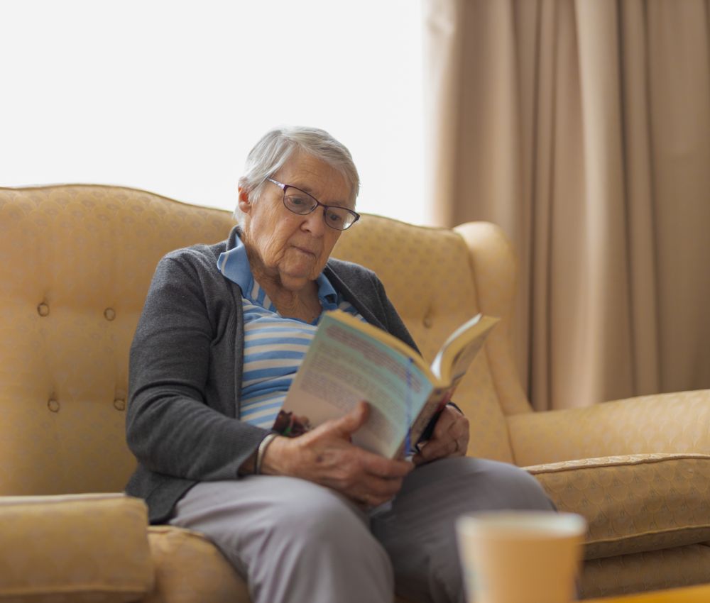 An Eldercare residents sits in a sun-soaked room while sitting in an armchair and reading a book.