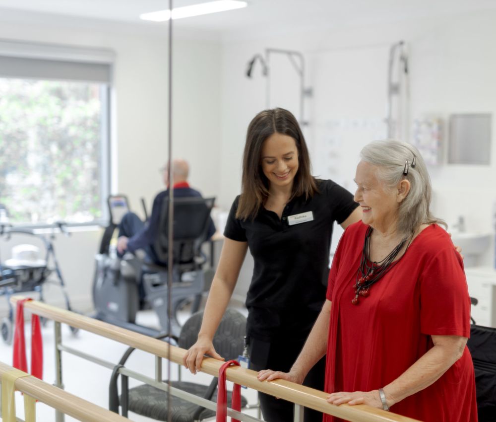 An Eldercare physiotherapist stands with a resident at a set of parallel bars in the gym at Allambi.