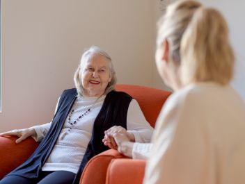 An Eldercare resident smiles at her daughter and holds her hand while sitting in an armchair.