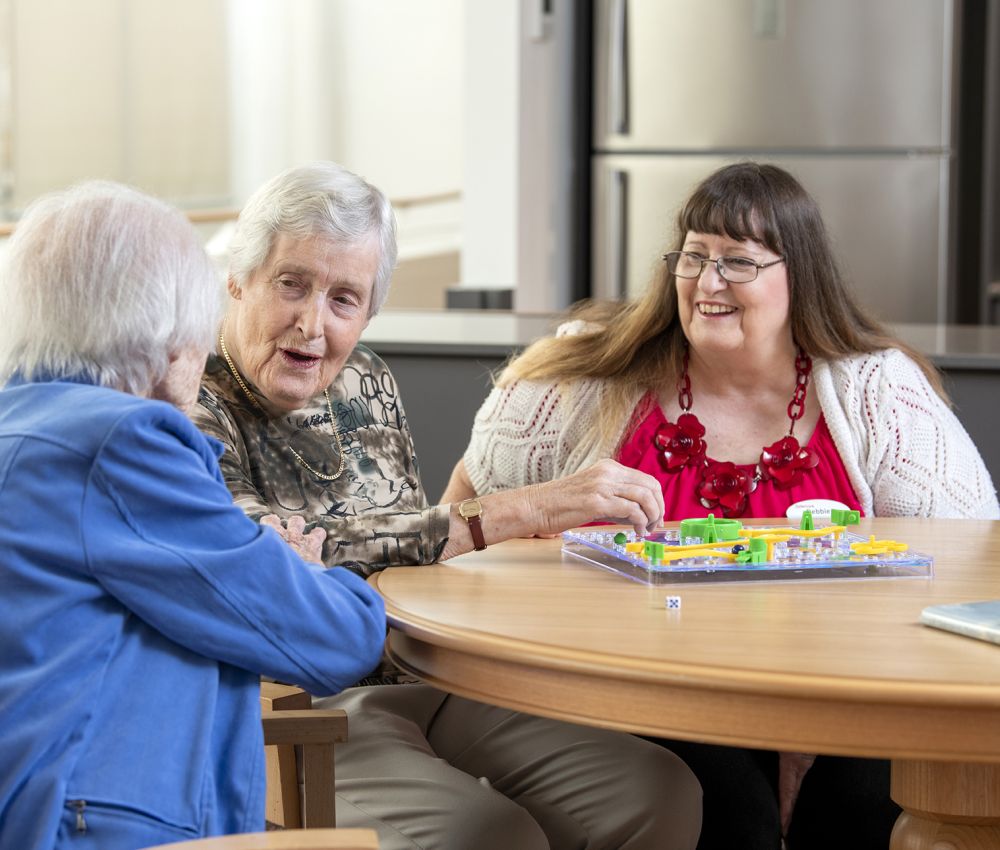 An Eldercare volunteer watches as two residents chat while sitting in front of a board game.