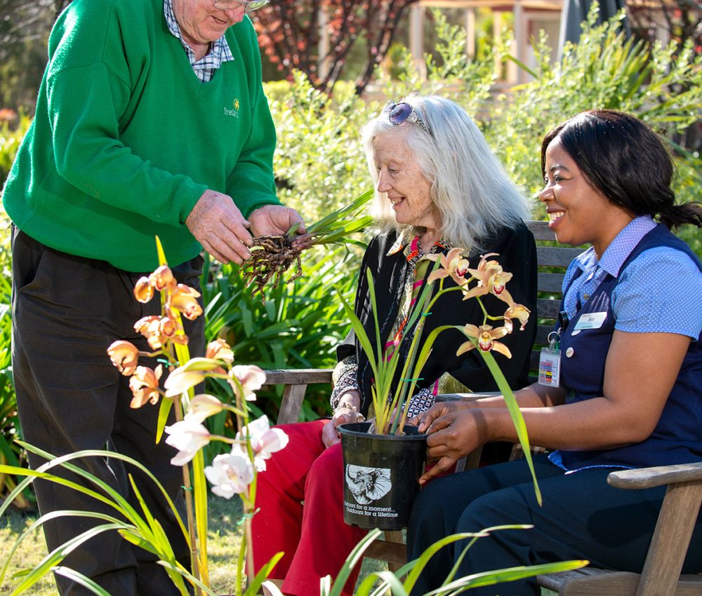 An Eldercare resident sits outdoors with a carer while a volunteer stands next to them and shows them some flower bulbs.