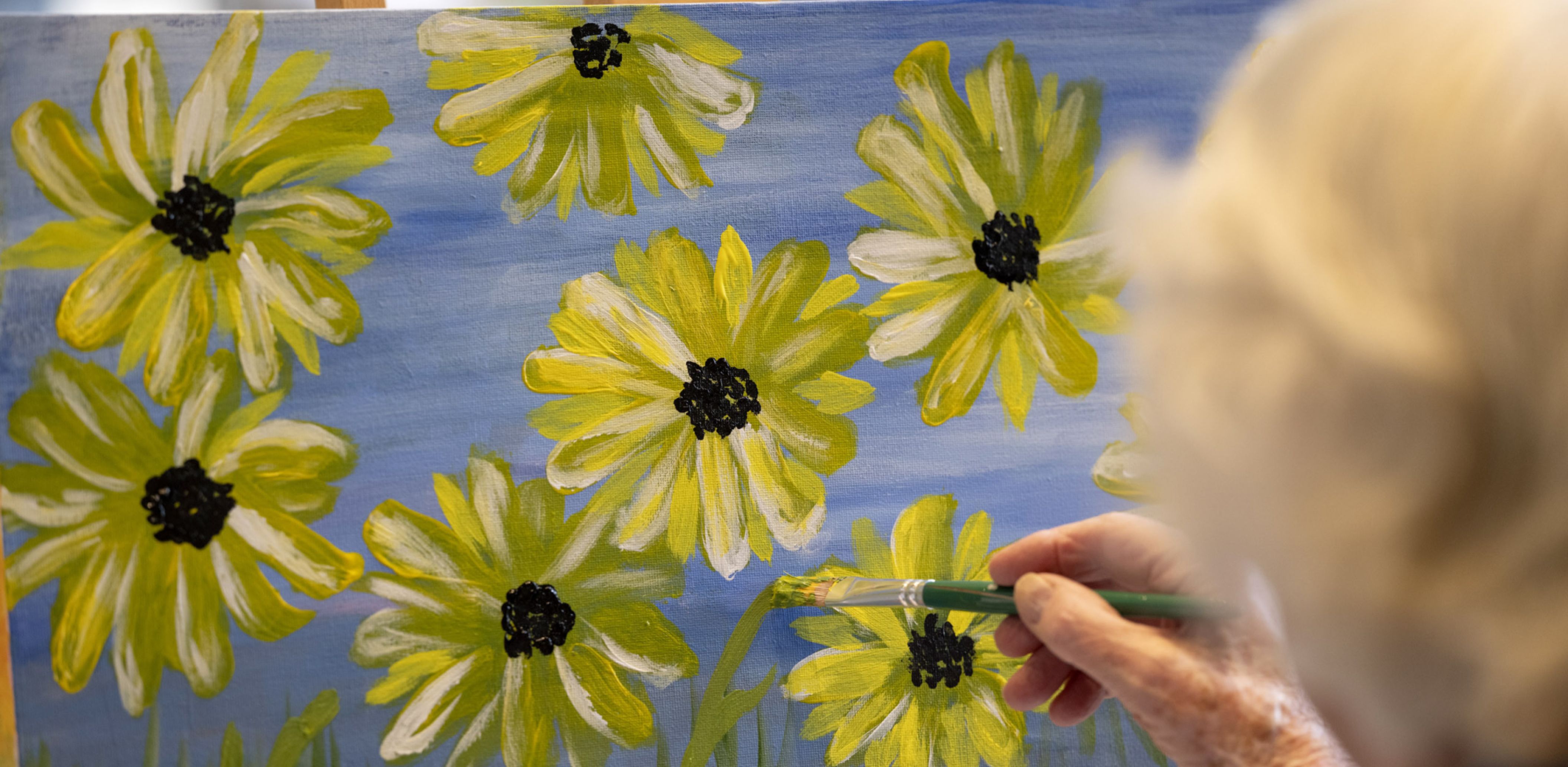 Eldercare resident paints a canvas featuring vibrant yellow flowers in a bright, sunlit space.
