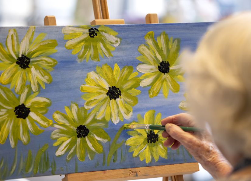 Eldercare resident paints a canvas featuring vibrant yellow flowers in a bright, sunlit space.