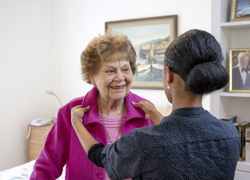 An Eldercare carer assists a resident in putting on her jacket, showcasing a moment of care.