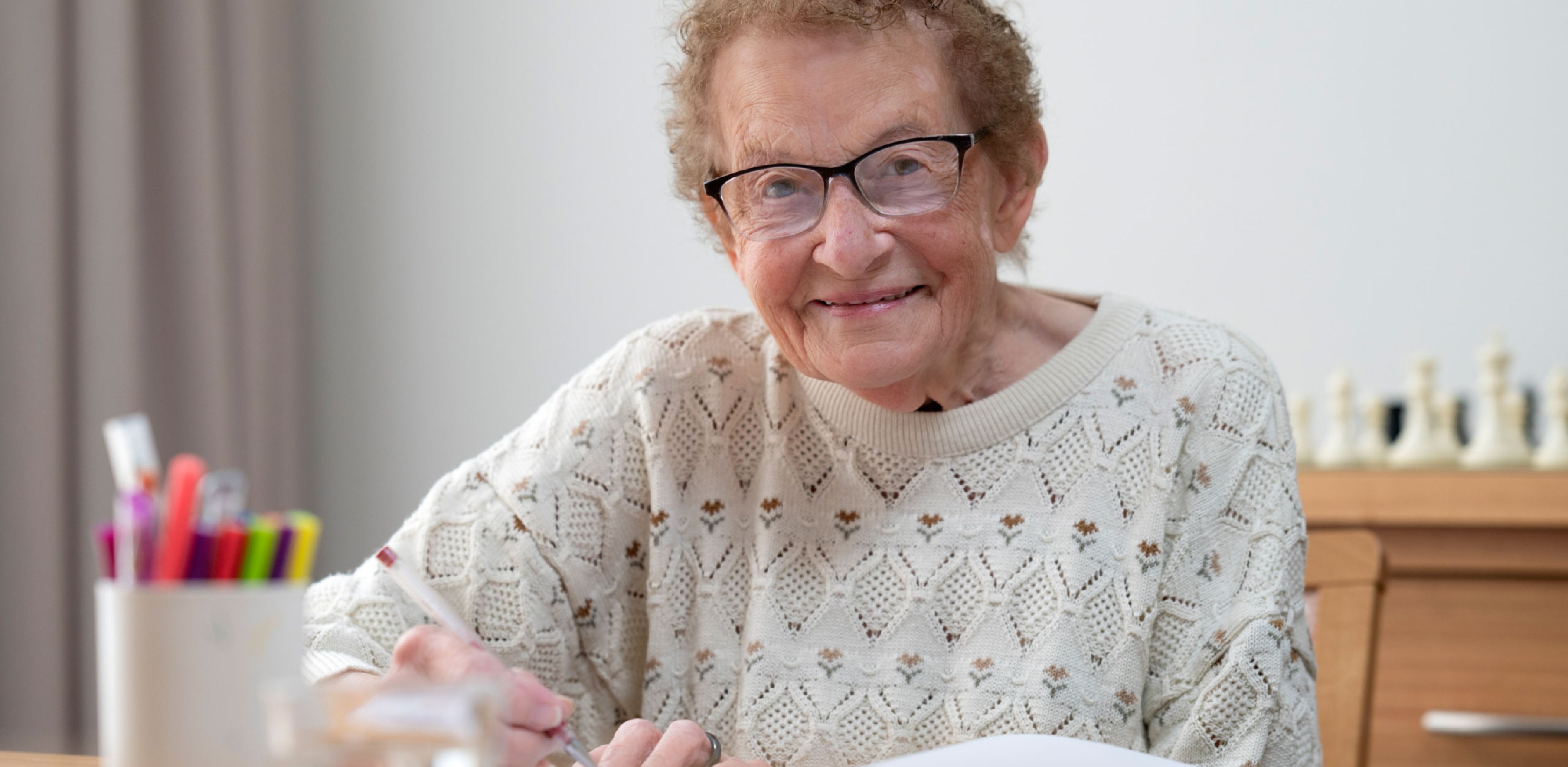 An Eldercare resident in a sweater and glasses writes in a book.