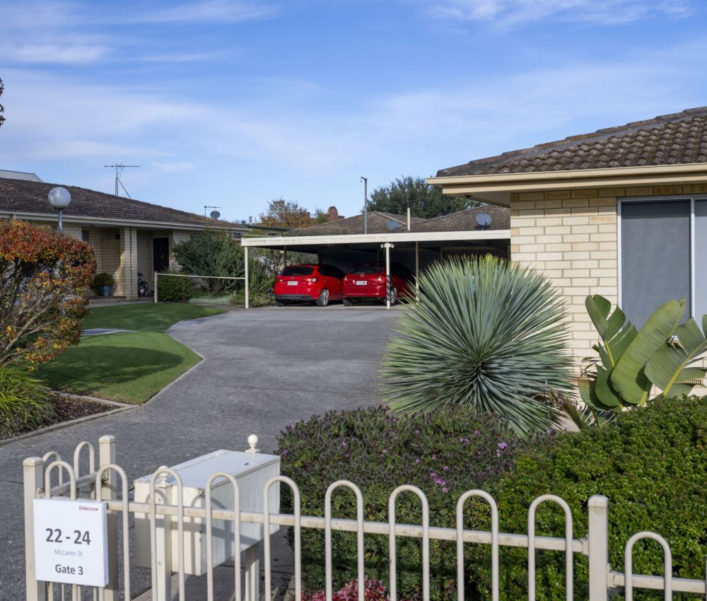 Well-maintained unit complex with cream brick buildings, landscaped gardens, gated entry, and driveway leading to covered carports with parked vehicles.