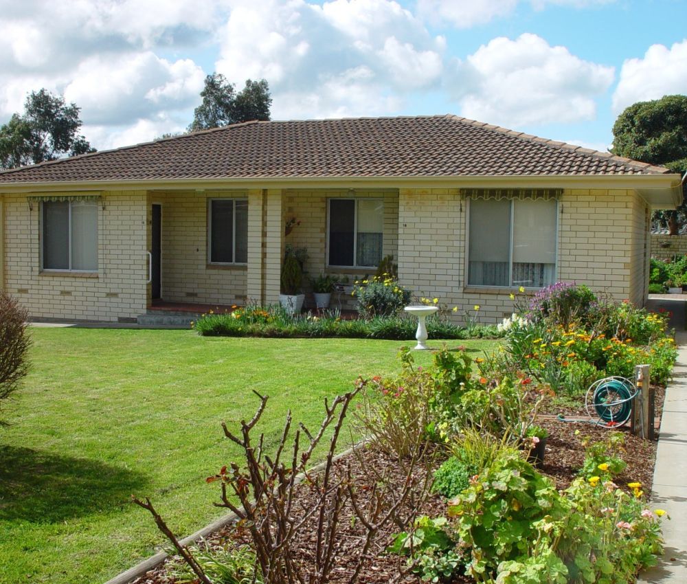 Cream brick single-level house with tiled roof, large front lawn, colourful garden beds, pathway and well-kept landscaping under a bright sky.