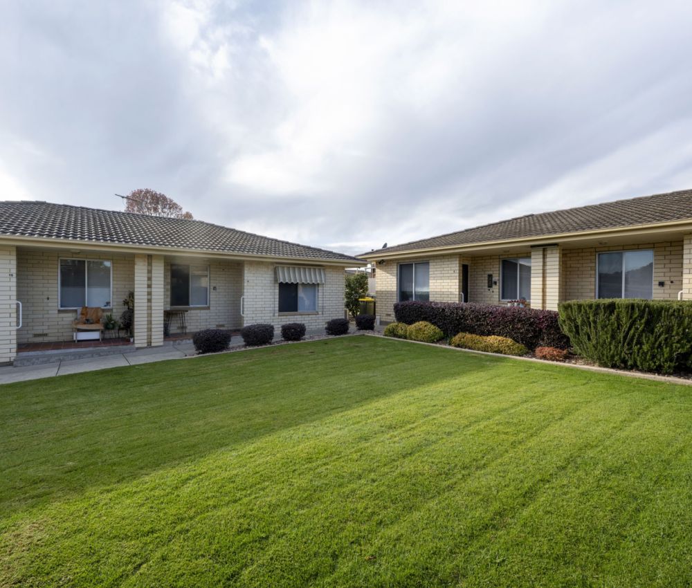 Spacious shared courtyard with neatly maintained lawn, surrounded by single-storey cream brick units and landscaped garden beds under a cloudy sky.