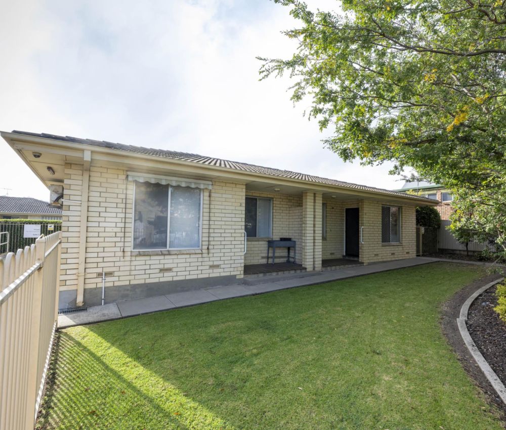 Single-storey cream brick house with tiled roof, front lawn, fenced yard and covered entry, surrounded by mature trees and garden beds.