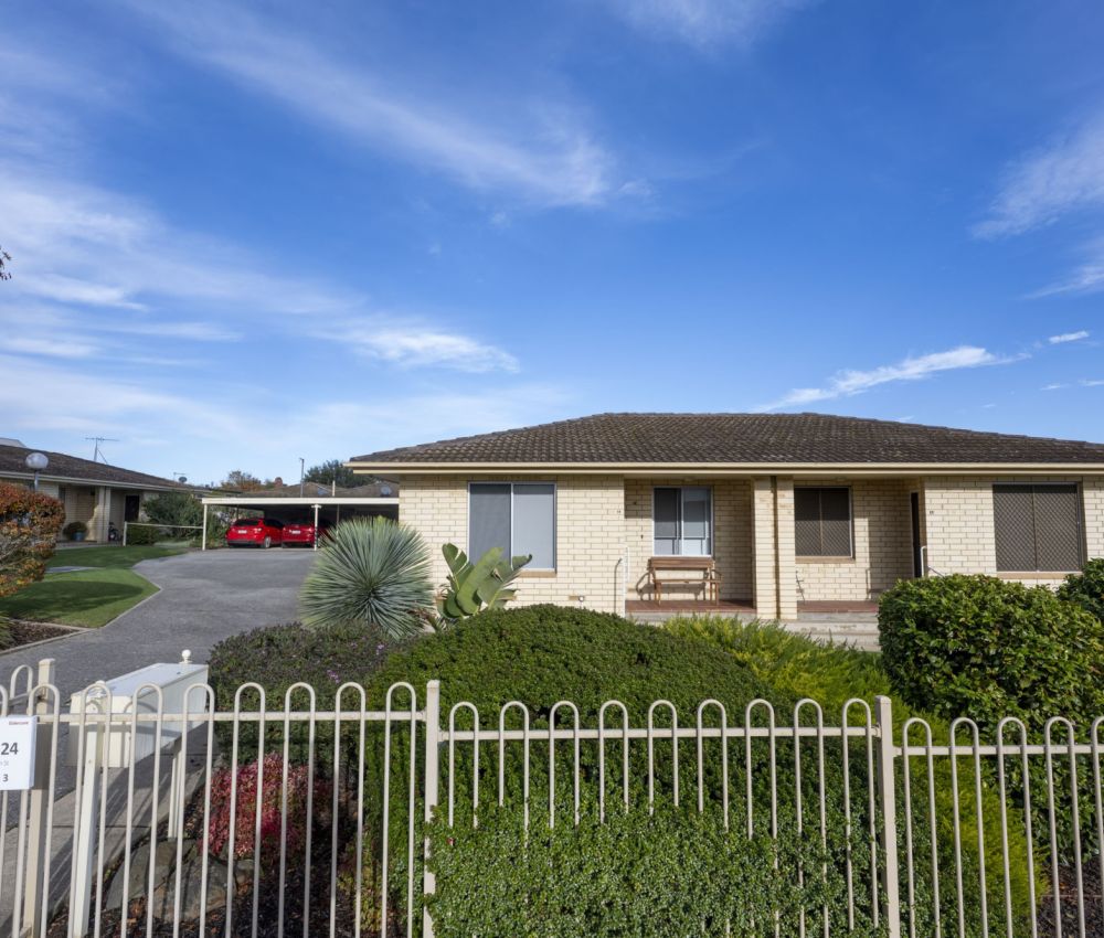 Single-storey cream brick home with tiled roof, enclosed front garden, white fence, driveway access and neighbouring units under a blue sky.