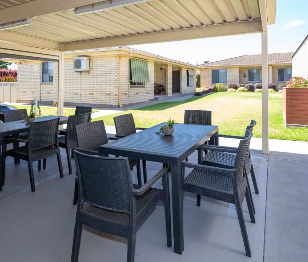 Covered outdoor patio with dining tables and chairs overlooking a spacious backyard lawn and single-storey brick home.