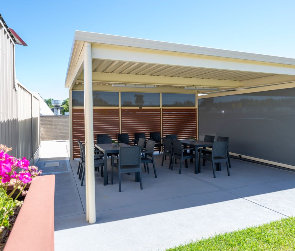 Modern outdoor entertaining area with covered pergola, dining tables and chairs on a paved patio, privacy screens, and adjacent lawn with garden flowers.