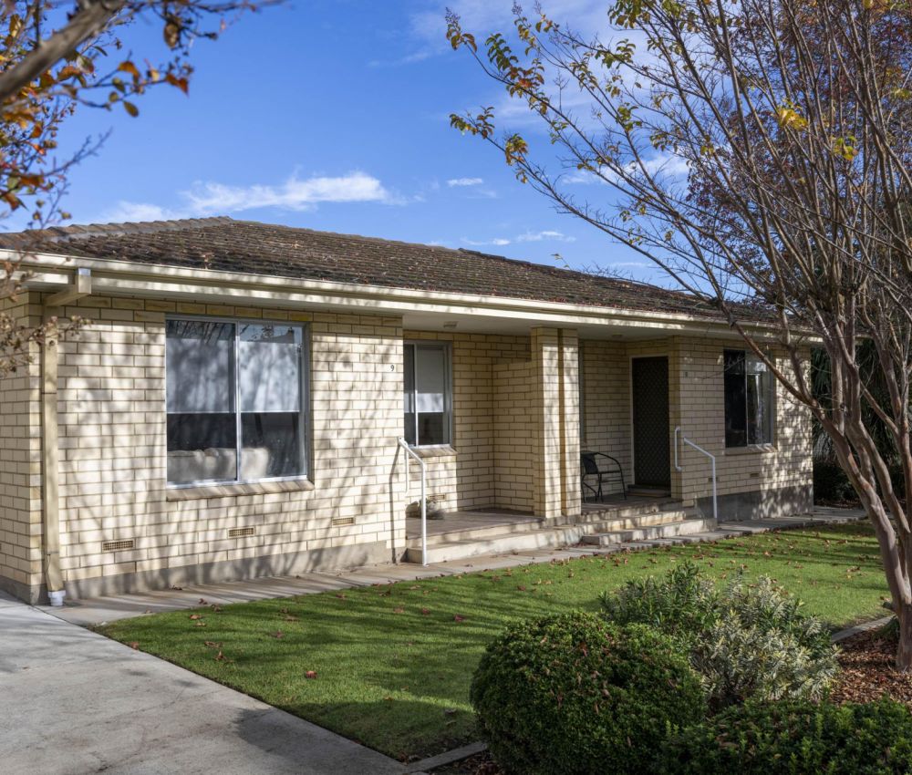 Single-storey cream brick home with covered front porch, driveway access, neat lawn and landscaped garden, framed by trees in a quiet residential setting.