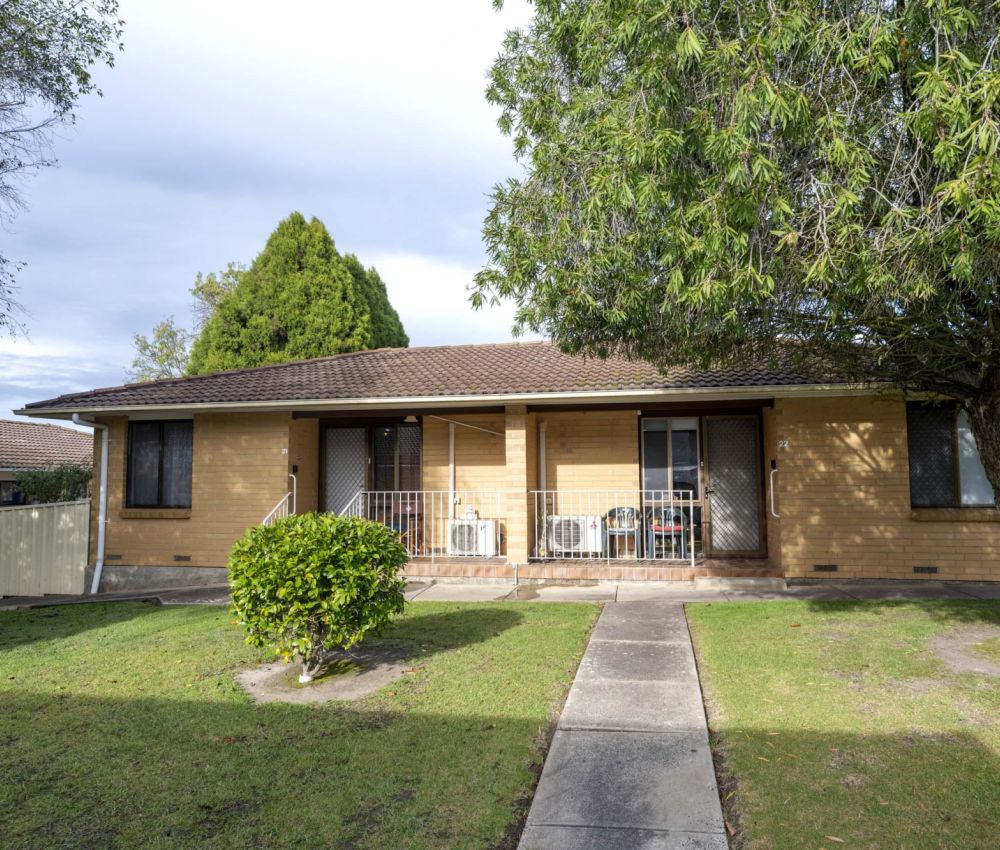 Single-storey cream brick unit with front porch seating, central pathway, lawn and mature tree, set within a quiet, well-maintained residential complex.