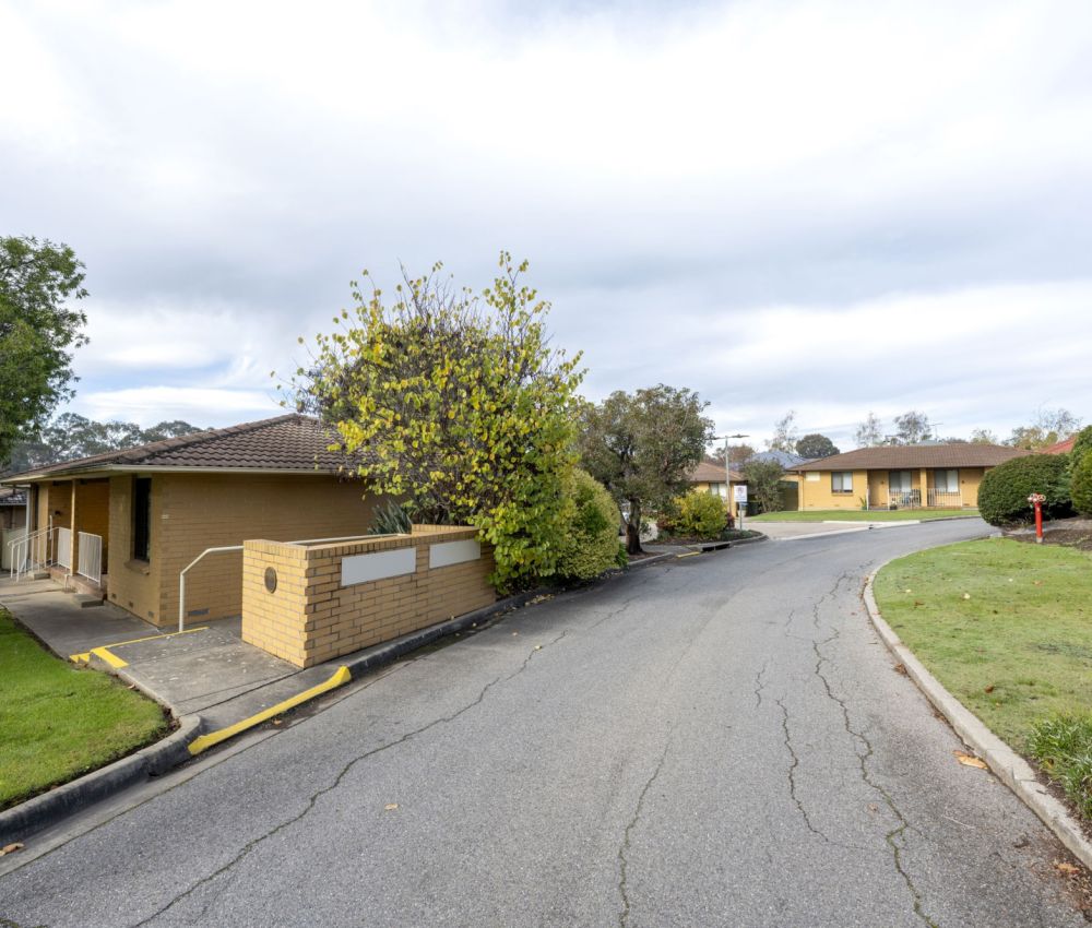 Internal street view of a well-maintained unit complex featuring cream brick buildings, landscaped gardens, curved driveway, and neatly trimmed lawns under an overcast sky.