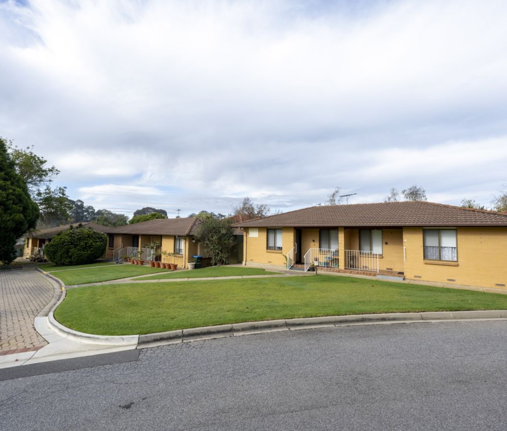 Curved street view of a single-storey cream brick unit complex with tiled roofs, neatly maintained lawns, paved driveway, and landscaped surroundings under a cloudy sky.