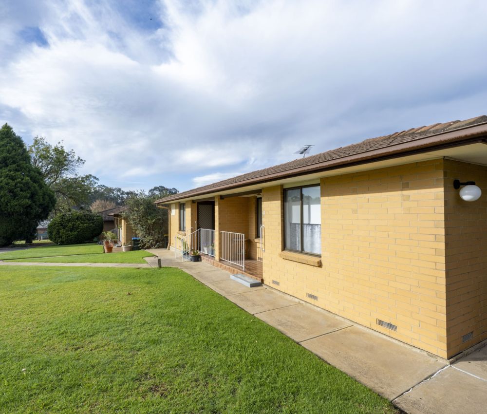 Single-storey cream brick unit with tiled roof, front porch with railing, concrete pathway, and wide green lawn within a well-kept residential complex.