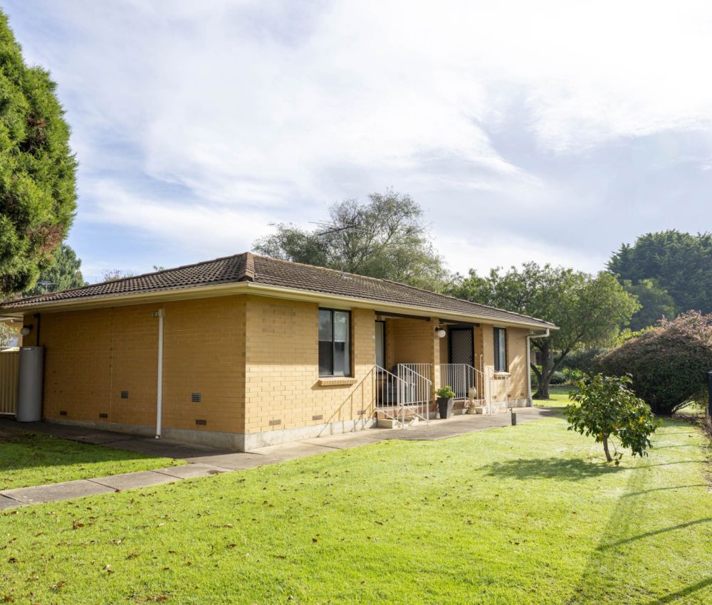 Single-storey cream brick home with side lawn, paved pathway, front steps with railing, and surrounding trees and garden in a quiet residential setting.