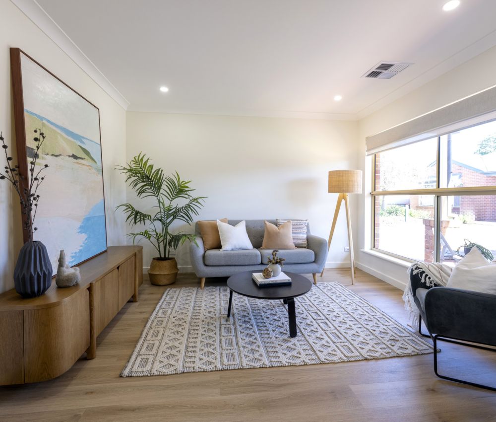 Bright, modern living room with a sofa, armchair, coffee table, large window, indoor plant, and wooden cabinet.