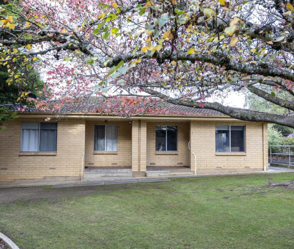 Single-storey cream brick unit with central porch, lawn and paved walkway, framed by a large tree with autumn leaves, set within a quiet and well-maintained residential complex.