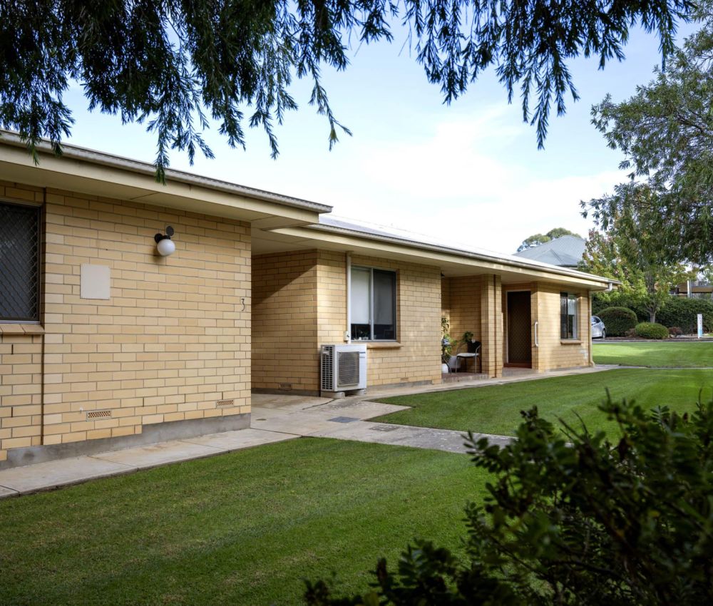 Row of single-storey cream brick units with covered walkways, air conditioning units, and neatly maintained lawns, partially framed by trees and shrubs in a quiet residential setting.